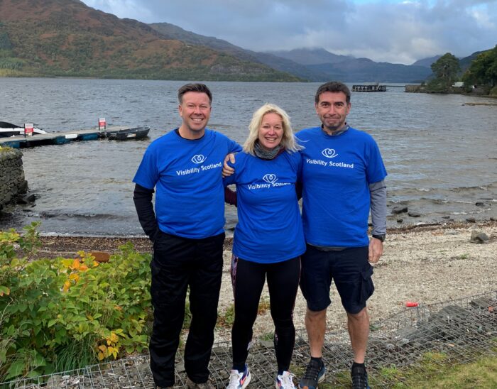 Sarah, with husband Andy on the right and friend Steven on the left. They are wearing Visibility Scotland t-shirts and standing in front of a loch after completing their three peaks challenge.