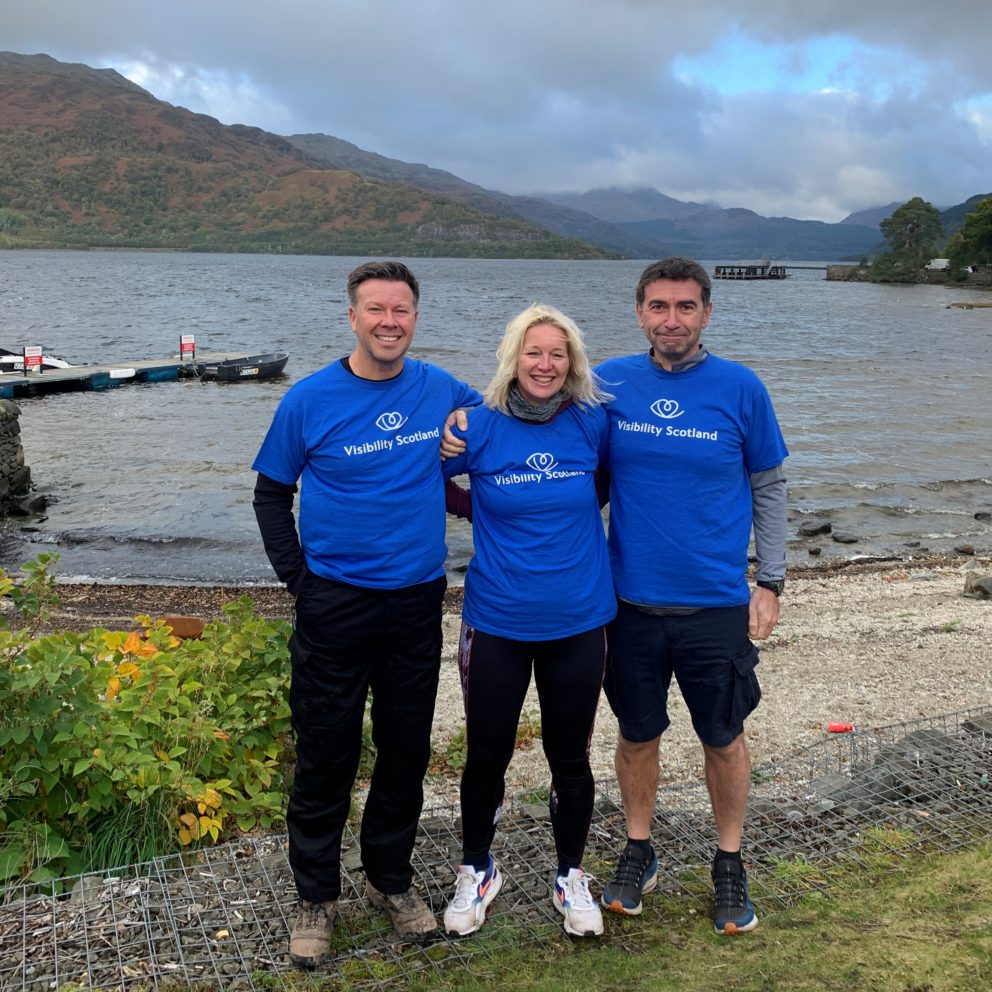 Sarah, with husband Andy on the right and friend Steven on the left. They are wearing Visibility Scotland t-shirts and standing in front of a loch after completing their three peaks challenge.