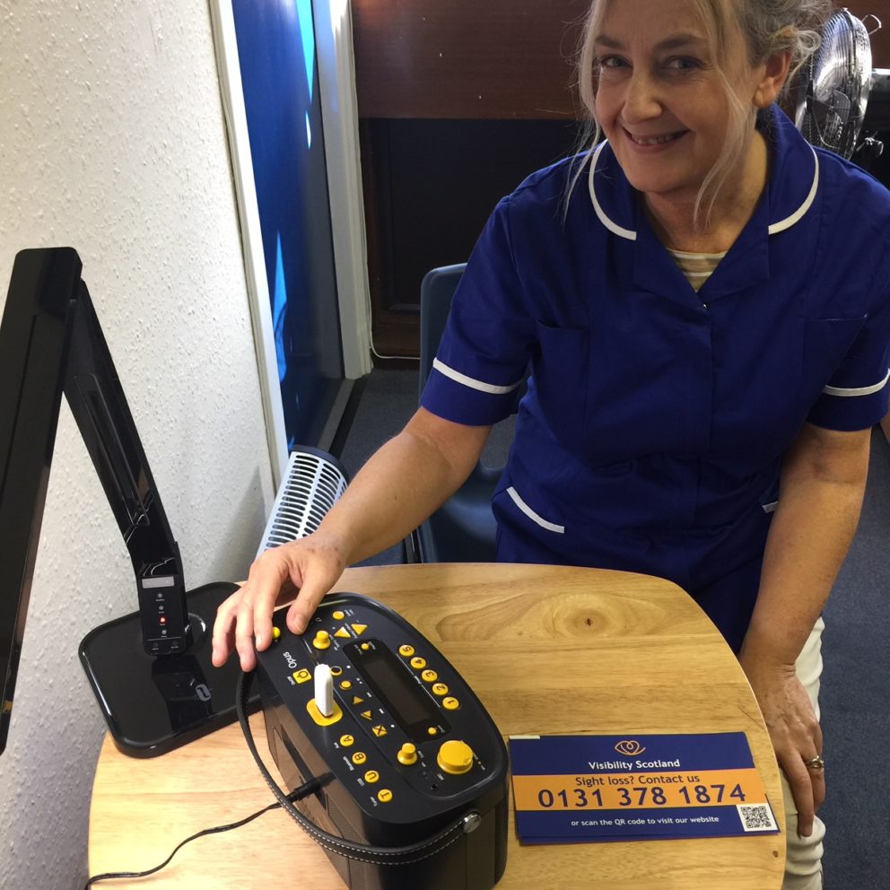 Visibility Scotland staff member, Jo, in our Patient Support room at the Princess Alexandra Eye Pavilion. Jo is sitting at a table and has a task lamp and big button radio on display to show patients.