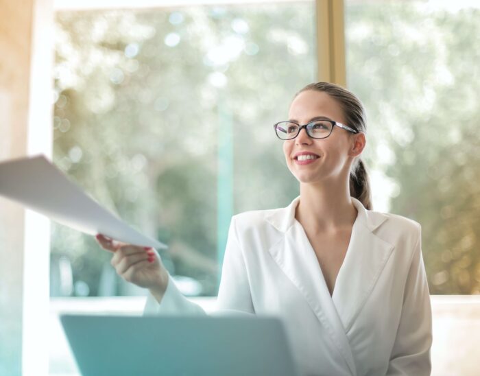 A woman sitting at a desk wearing a white shirt and glasses reaching out to receive paperwork from a person out of frame