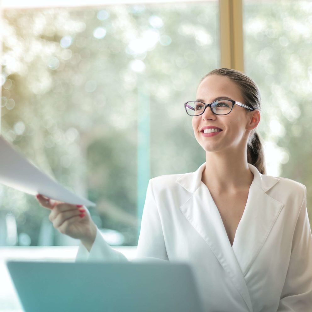 A woman sitting at a desk wearing a white shirt and glasses reaching out to receive paperwork from a person out of frame