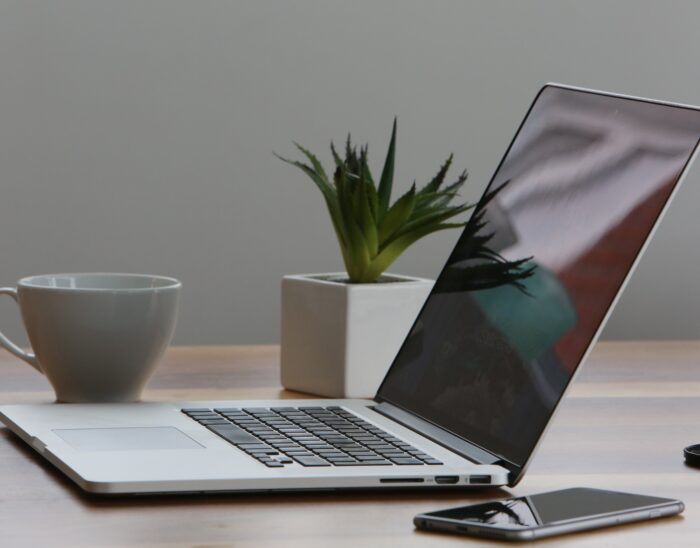A laptop and smartphone sitting on a desk next to a pot plant and coffee cup