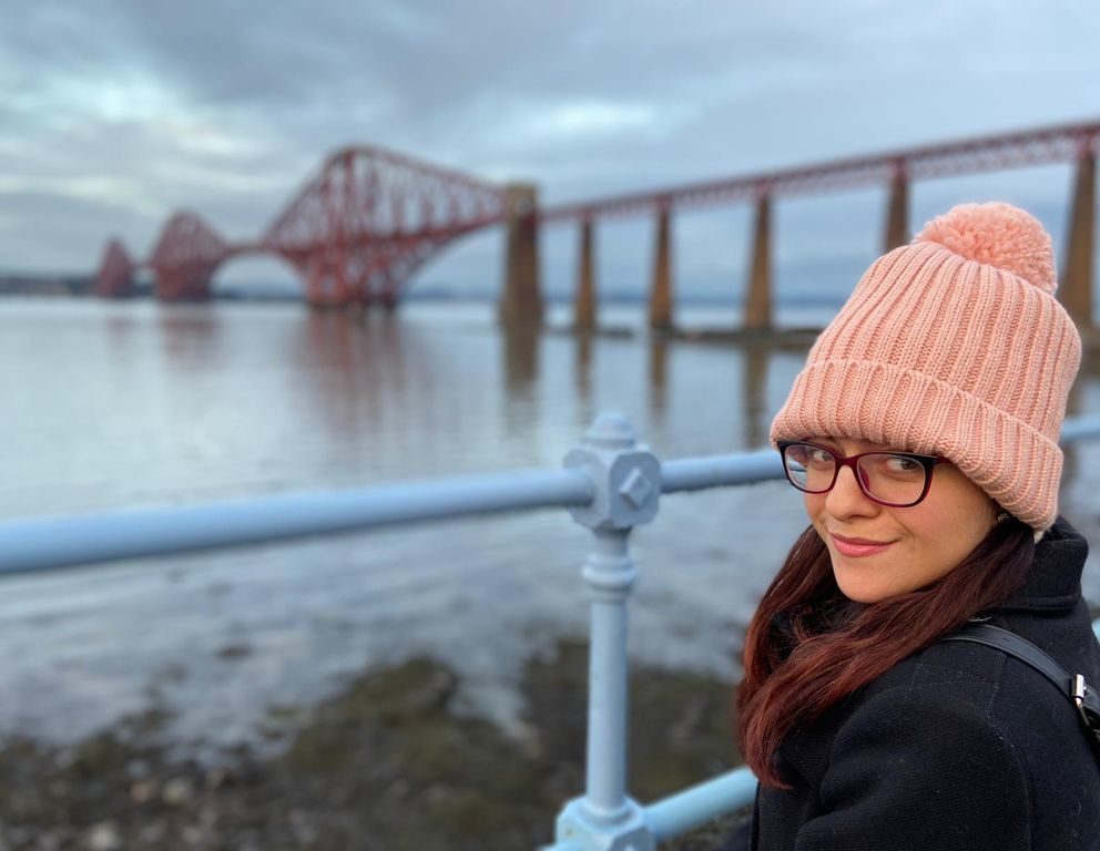 Zee wearing a pink bobble hat, standing at railings with the Forth rail bridge in the background