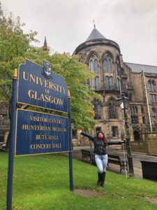 Zein standing outside the University of Glasgow. She has her arms spread wide and is holding her white cane, "Bonnie".