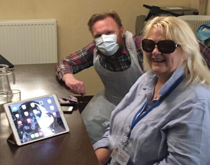 Nicola and Visibility Staff member, Craig, sitting at a table with an iPad and iPhone on it. Nicola is wearing dark glasses and Craig is wearing PPE.