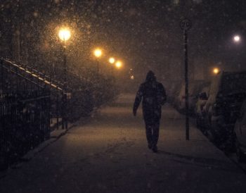 A person walking along a snow-covered pavement at night. Snow is falling and the image is dark despite some lighting on the path.