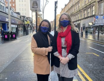 Laura and Audrey wearing Visibility Scotland facemasks, standing on Sauchiehall Street. Audrey is holding her long cane.
