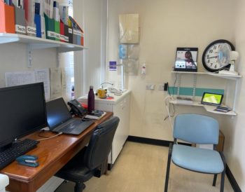 The Patient Support room in Gartnavel eye department. There are example aids on display (high contrast, large number clock, big button phone, liquid level indicator etc.), a computer, laptop and phone on a desk and folders of information on a self.