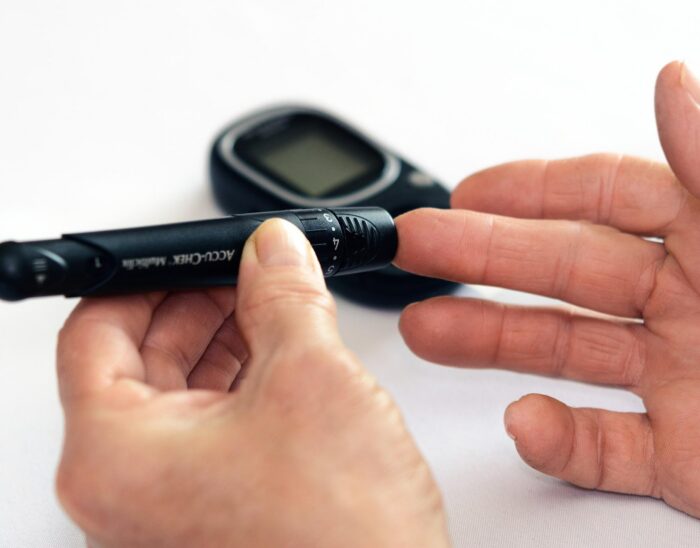 Close up of a person's hands whilst they carry out a pinprick test to check their blood sugar levels. A glucose meter is visible in the background.