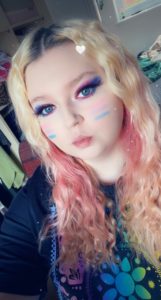 Erin ready for Pride in a black t-shirt with rainbow mandela pattern and the bisexual pride flag on her cheeks which starts with pink then purple then ends with blue and the same colours on her eyelids.