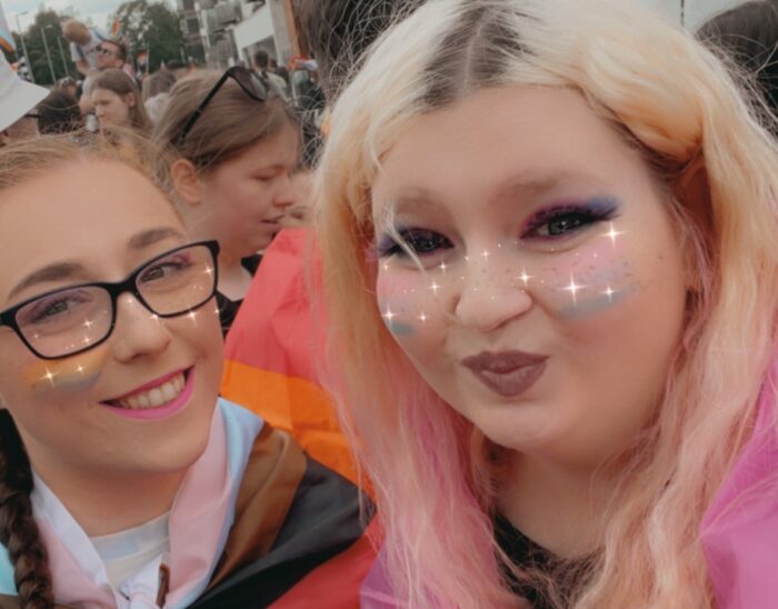Erin and a friend at pride with their flags. Erin's friend has the ally flag which consists of a black and white striped background and a rainbow triangle in the middle. Erin has the bisexual flag with the following stripes: at the bottom is blue, then purple in the middle and at the top is pink. They have the flags wrapped around them so only a little bit of each flag is visible.