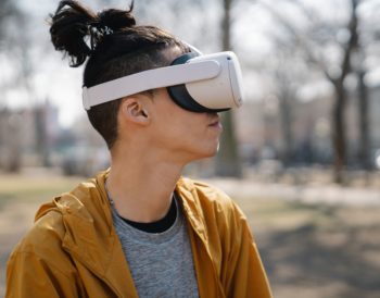 A person wearing a virtual reality headset. They are outdoors and their are trees in the background.