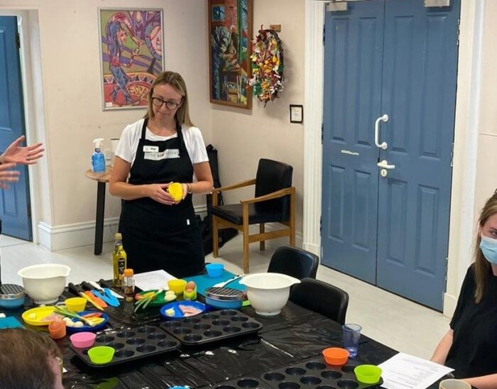 Jenn and Kay from Food Heroes leading a practical cookery session. Three EyeCan! participants are seated at a table. Various cooking aids are spread across the table including coloured chopping boards, silicone baking molds and high rim plates.