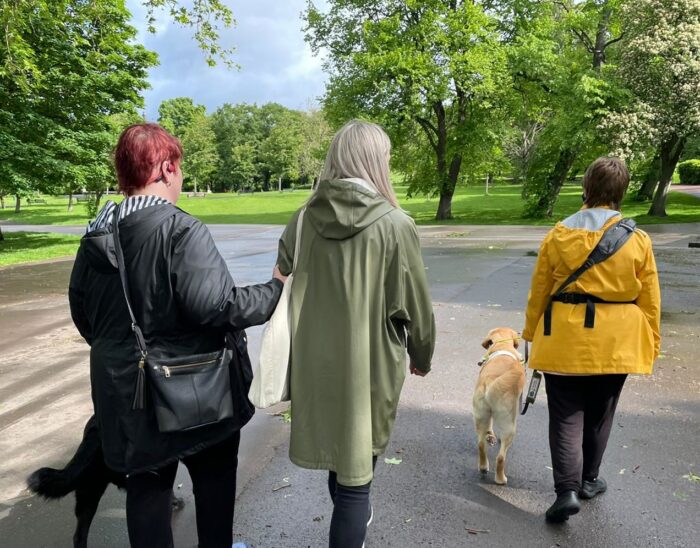 Three people walking in the park at our last Walk 'n' Talk event. Two of the walkers have guide dogs. A Visibility Scotland staff member is providing sighted guiding support to one of the walkers.