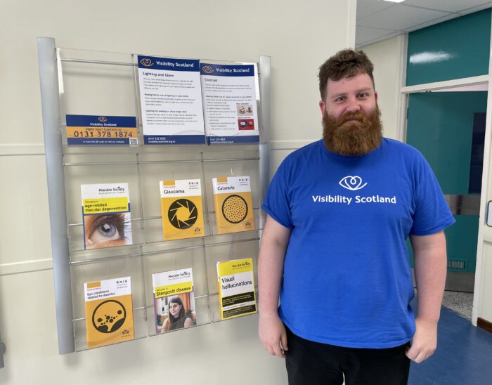 Callum wearing a Visibility Scotland t-shirt, standing next to a rack of information materials at the Princess Alexandra Eye Pavilion in Edinburgh.