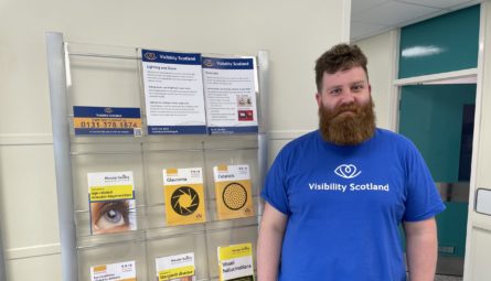Callum wearing a Visibility Scotland t-shirt, standing next to a rack of information materials at the Princess Alexandra Eye Pavilion in Edinburgh.