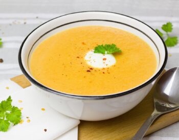 a bowl of carrot and coriander soup sits on a wooden board on a table.
