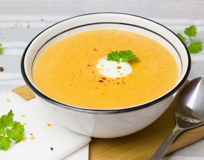 a bowl of carrot and coriander soup sits on a wooden board on a table.