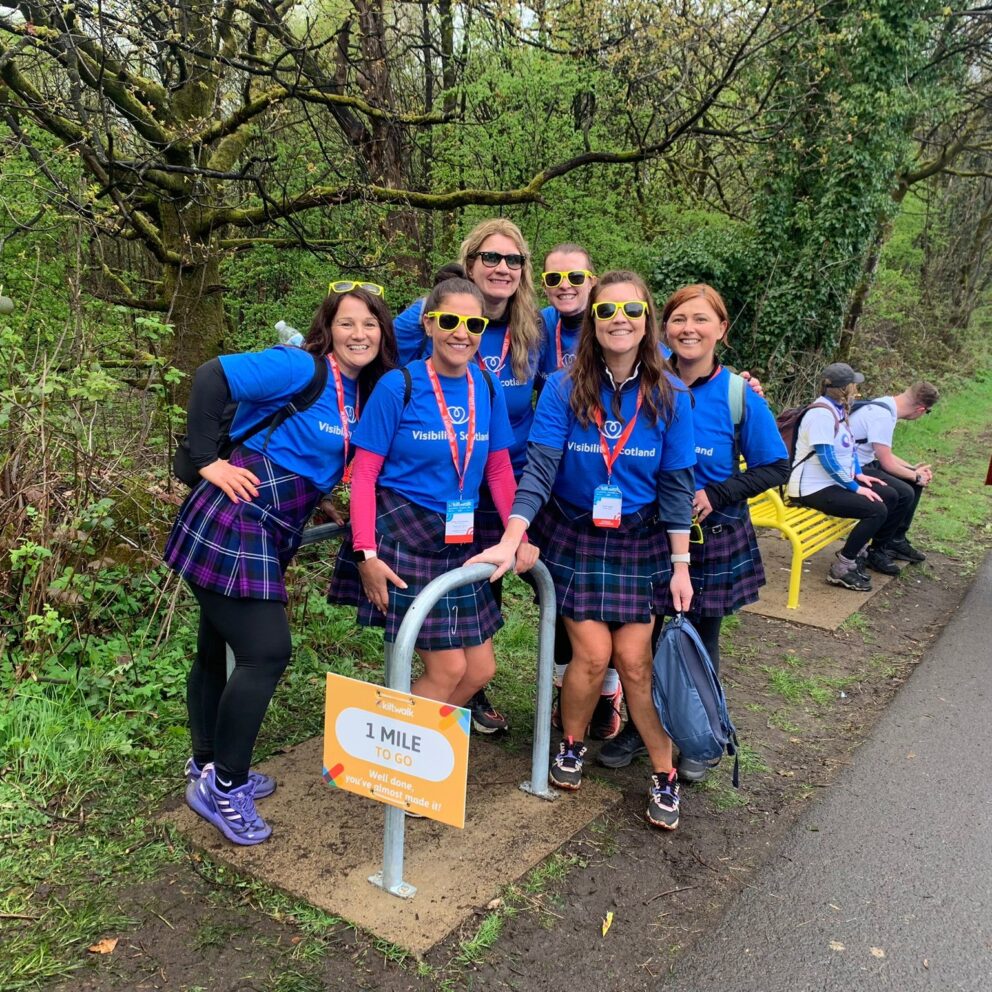 A group of Visibility Scotland fundraisers wearing our t-shirts and kilts, pose for a group photo at the Kiltwalk &#039;1 mile to go&#039; sign.