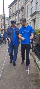 A visibility scotland staff member helps to guide another staff member who is wearing sleep shades and using a white long cane. They are walking along Queen's Crescent.