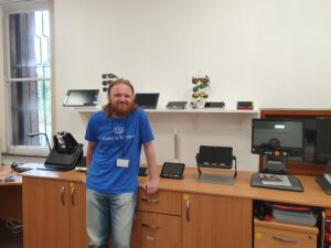 Craig smiles while wearing a blue Visibility Scotland-t-shirt in our digital suite. He is leaning against a table top with a range of digital magnifiers on it.