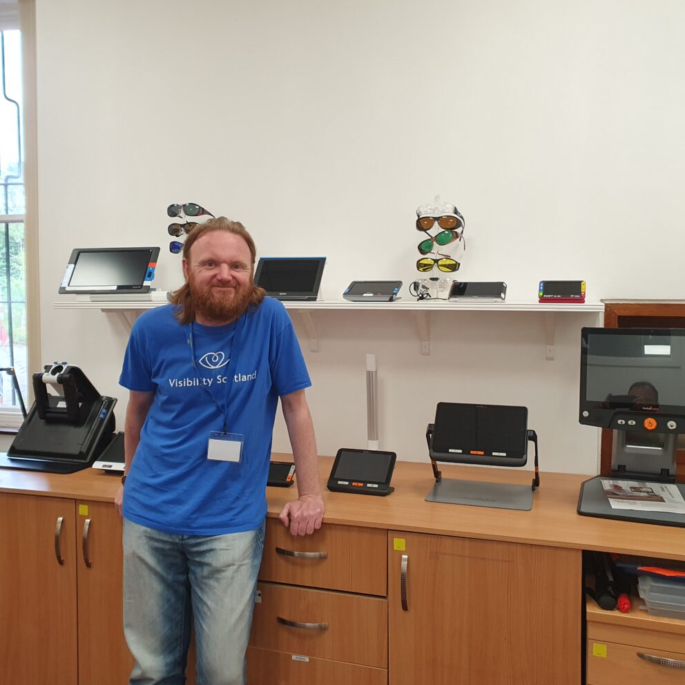 Craig smiles while wearing a blue Visibility Scotland-t-shirt in our digital suite. He is leaning against a table top with a range of digital magnifiers on it.