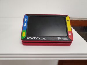 A Ruby XL HD digital magnifier. It has large colourful easy-to-see and tactile buttons against the black face of the device; the handle and outer casing are ruby red.