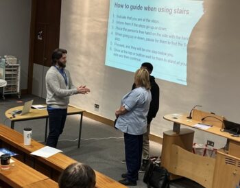 Adam stands in a lecture room in front of two people. Adam is explaining sighted guiding to them. The presentation slide projected on the wall explains how to guide someone when using stairs.