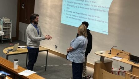 Adam stands in a lecture room in front of two people. Adam is explaining sighted guiding to them. The presentation slide projected on the wall explains how to guide someone when using stairs.