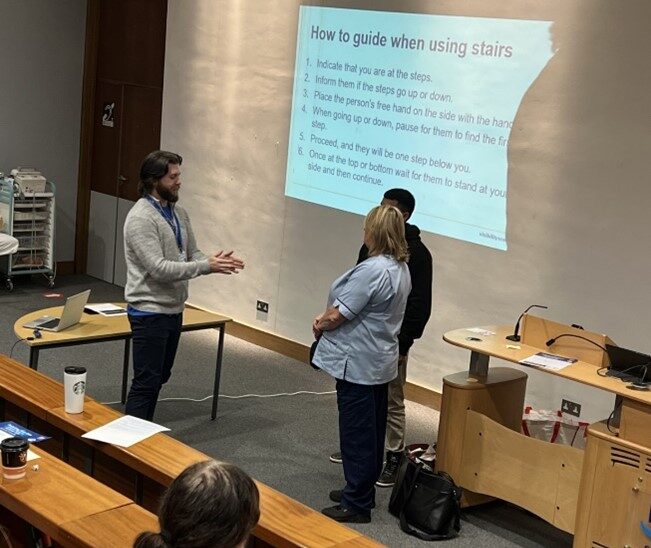 Adam stands in a lecture room in front of two people. Adam is explaining sighted guiding to them. The presentation slide projected on the wall explains how to guide someone when using stairs.
