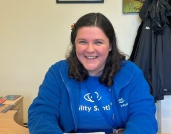 Erin sits at her desk in Mountainhall Treatment Centre. She is smiling while wearing a blue Visibility Scotland t-shirt and fleece