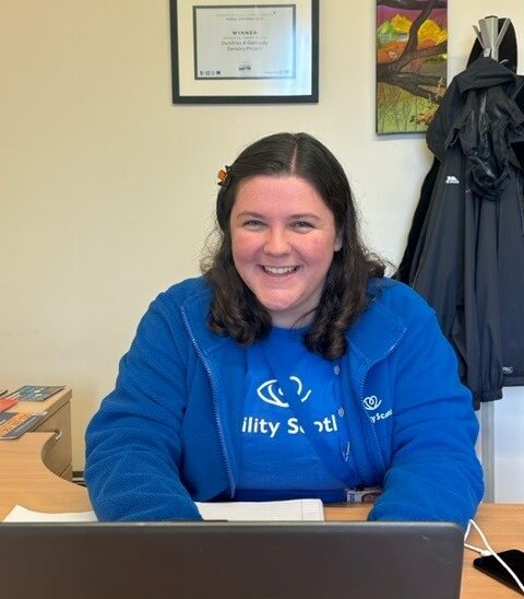 Erin sits at her desk in Mountainhall Treatment Centre. She is smiling while wearing a blue Visibility Scotland t-shirt and fleece