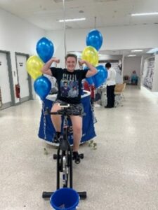One of the Boots fundraisers sits on a stationery bike in the Boots shop. She is smiling and flexing her arms in triumph.