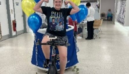 One of the Boots fundraisers sits on a stationery bike in the Boots shop. She is smiling and flexing her arms in triumph.