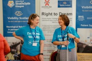 Claire and Audrey from Visibility Scotland stand in front of our information banner. They are wearing light blue concert for carers t-shirts