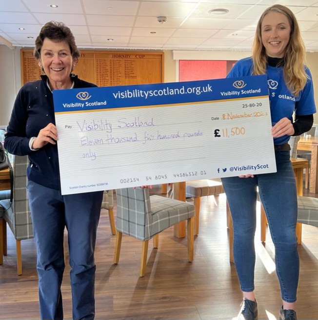 A female member of Cathcart Castle Golf Club smiles while holding one end of a presentation cheque for £11,500 made out to Visibility Scotland. Laura Walker, CEO of Visibility, smiles while holding the other end.