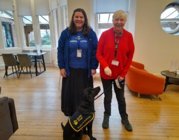 Erin, Visibility Scotland&#039;s community worker, stands smiling alongside a volunteer from Guide Dogs, who has a black Labrador sitting on the floor on a lead. Erin and the volunteer are smiling.
