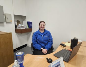 August sits behind the reception desk. She is smiling and is wearing a blue Visibility Scotland fleece jacket.