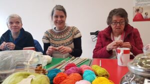 Three people sit knitting at a table full of brightly coloured yarn