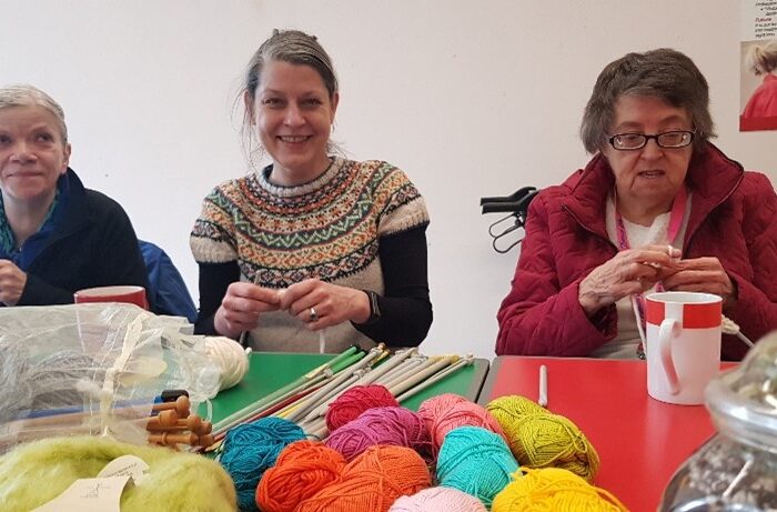 Three people sit knitting at a table full of brightly coloured yarn
