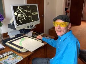 Photo of Roy sitting at his desk with his digital magnifier in front of him showing a map of the stars. He is smiling.