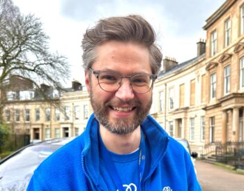 Alister Lees smiles while standing outside our main office. he is wearing a blue Visibility Scotland t-shirt and fleece. Ali wears glasses and has a beard.