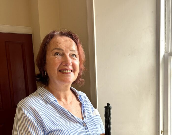 Audrey is standing next to a window wearing a blue and white striped shirt and holding her guide cane. She is smiling.