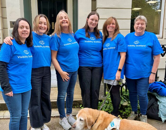 Visibility Scotland's patient support team stands together smiling outside the main office building. Gina, a yellow lab, stands in front of the group.