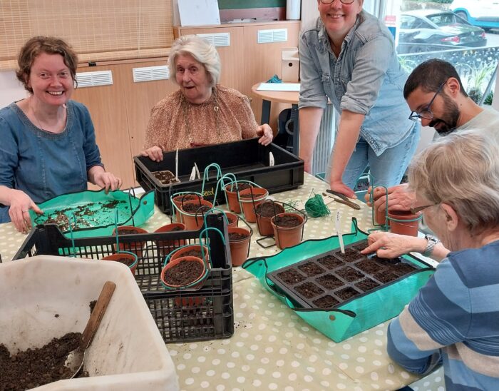 Four people sit around a table planting seeds, another person stands beside the table smiling.