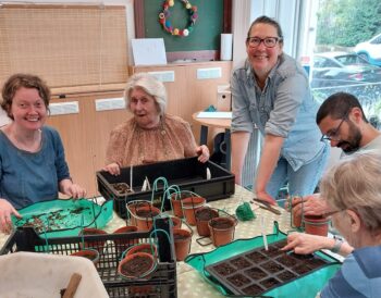 Four people sit around a table planting seeds, another person stands beside the table smiling.