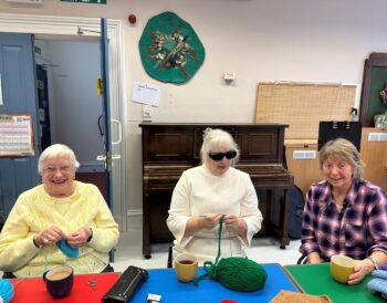 Three people sit at a table smiling. Two people are knitting and one is holding a mug.
