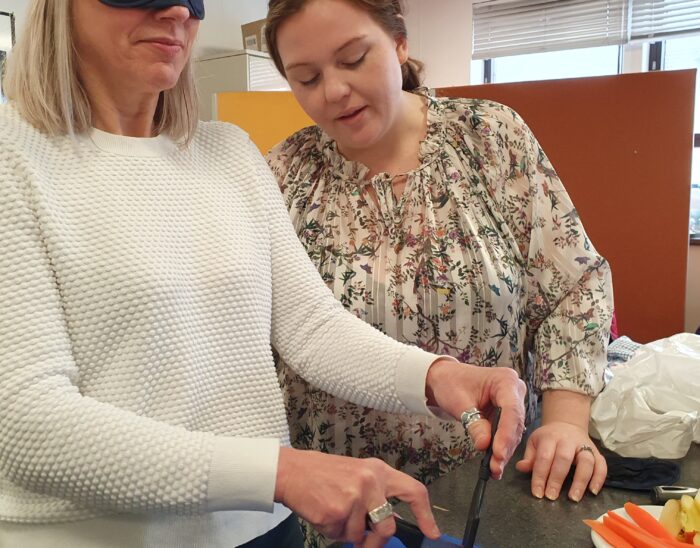 A person in a blindfold cuts vegetables while another person carefully observes.