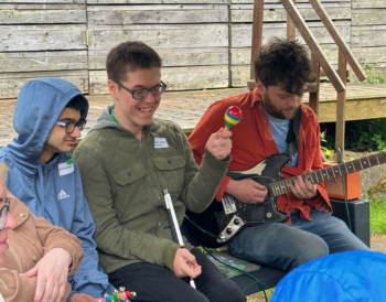 Three people sit together playing musical instruments and smiling