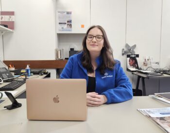 Lydia smiles while seated at her laptop. She has long brown hair, glasses and is wearing a blue Visibility Scotland fleece jacket